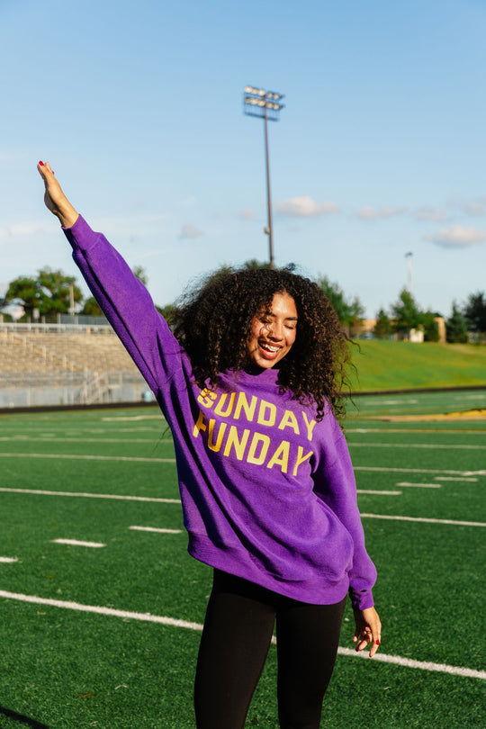 A smiling person with curly hair stands on a track field, raising one arm and posing cheerfully in the sunlight while wearing the Panache Apparel Collection Purple Game Day & Sunday Funday Reversible Sweatshirt.
