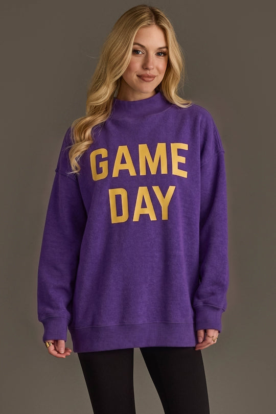 A woman with long blonde hair wears the Panache Apparel Collection Purple Game Day & Sunday Funday Reversible Sweatshirt, featuring bold yellow “GAME DAY” lettering, paired with black leggings and standing against a plain gray background.