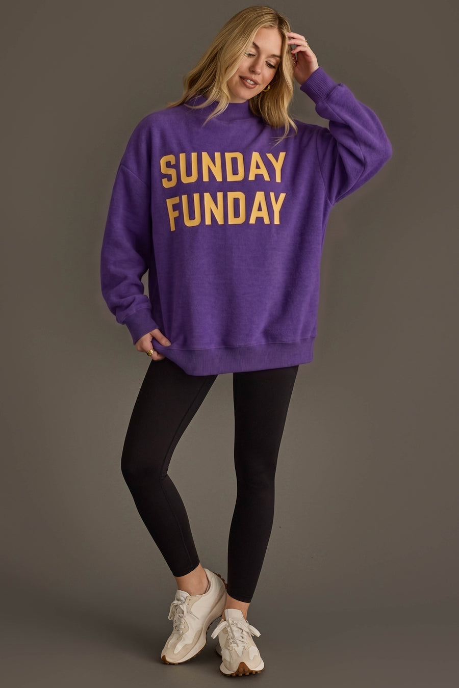 A woman stands smiling, wearing the Panache Apparel Collection Purple Game Day & Sunday Funday Reversible Sweatshirt featuring bold yellow text, paired with black leggings and white sneakers, against a plain brown background.