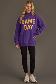 A woman stands smiling in a Panache Apparel Collection Purple Game Day & Sunday Funday Reversible Sweatshirt featuring yellow “GAME DAY” lettering, paired with black leggings and white sneakers, set against a plain gray background.