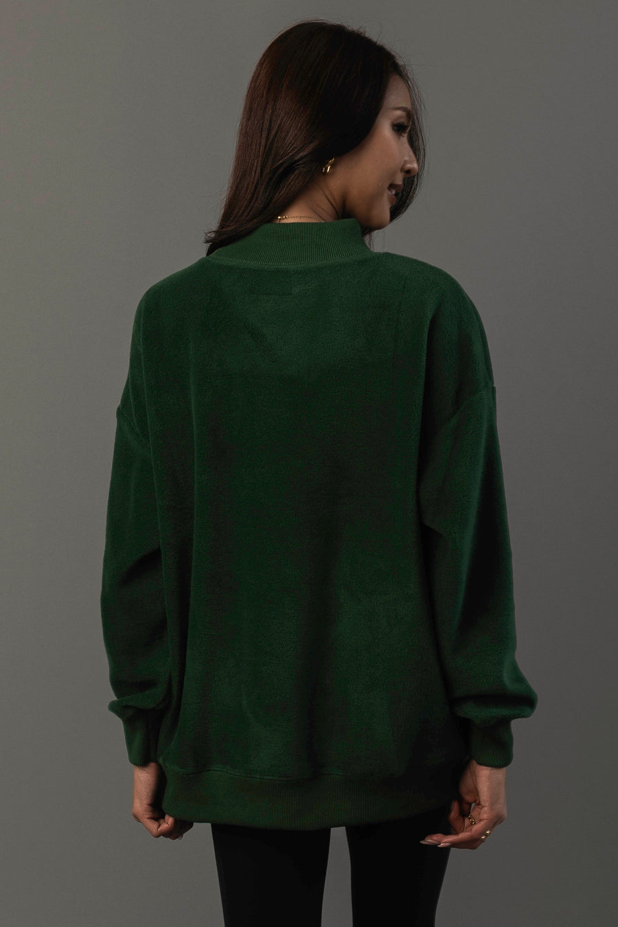 A woman with long dark hair, wearing the Panache Apparel Collection’s Green Game Day & Sunday Funday Reversible Sweatshirt and black pants, stands facing away from the camera against a plain gray background.