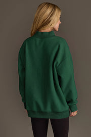 A person with long blonde hair is standing with their back to the camera, wearing the Panache Apparel Collection Green Game Day & Sunday Funday Reversible Sweatshirt and black pants, against a plain brown background.