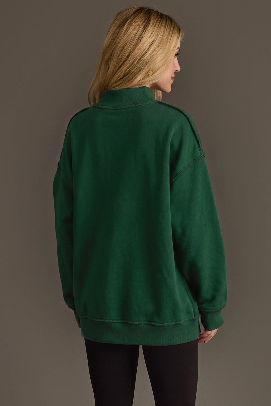 A person with blonde hair, wearing the Panache Apparel Collection Green Game Day & Sunday Funday Reversible Sweatshirt and black leggings, stands facing away from the camera against a plain brown background.