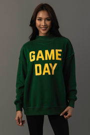 A woman stands and smiles against a plain gray background, wearing the Panache Apparel Collection’s Green Game Day & Sunday Funday Reversible Sweatshirt with bold yellow “GAME DAY” lettering.