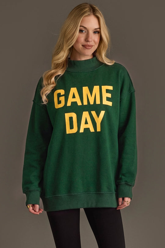 A woman with long blonde hair wears the Panache Apparel Collection’s Green Game Day & Sunday Funday Reversible Sweatshirt—a vibrant green top featuring bold yellow “GAME DAY” lettering. She stands against a plain background and smiles slightly.