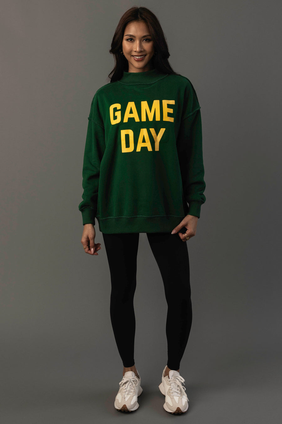 A woman stands smiling against a plain gray background, wearing the Panache Apparel Collection Green Game Day & Sunday Funday Reversible Sweatshirt, paired with black leggings and white sneakers.