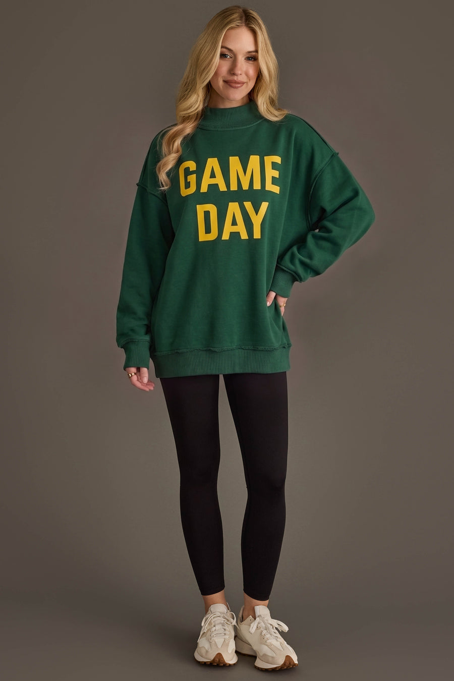 A woman stands smiling, wearing the Green Game Day & Sunday Funday Reversible Sweatshirt by Panache Apparel Collection paired with black leggings and white sneakers, against a plain gray background.