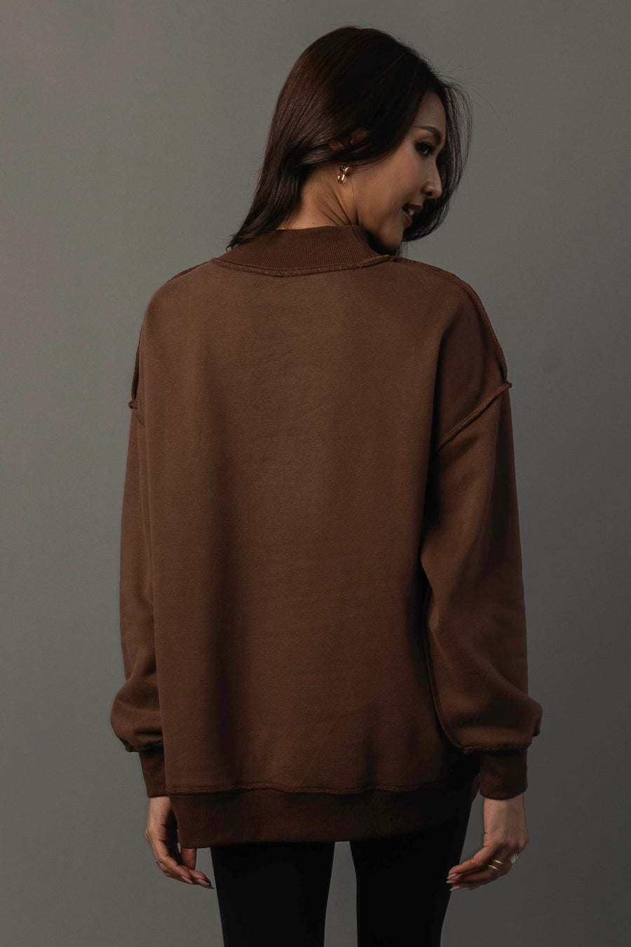 A woman with long brown hair, standing with her back to the camera and turning her head to the side against a neutral gray background, models the Panache Apparel Collection’s Brown Whiskey Weather & Bourbon Weather Reversible Sweatshirt.
