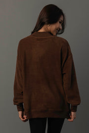 A woman with long brown hair wears the Panache Apparel Collection’s Brown Whiskey Weather & Bourbon Weather Reversible Sweatshirt and black pants, standing with her back to the camera while looking over her shoulder and smiling against a plain gray background.