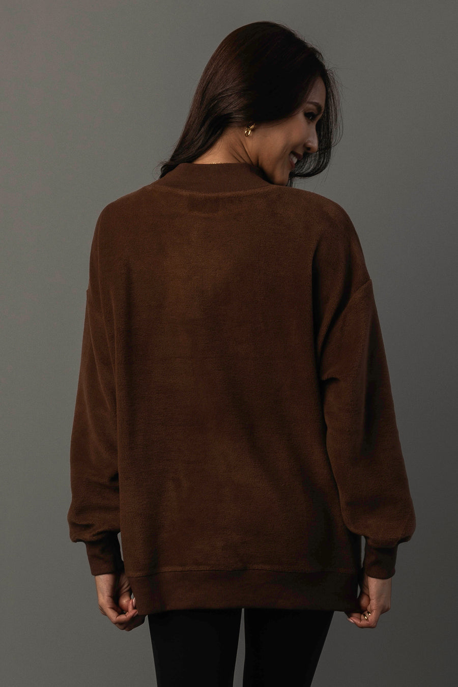 A woman with long brown hair wears the Panache Apparel Collection’s Brown Whiskey Weather & Bourbon Weather Reversible Sweatshirt and black pants, standing with her back to the camera while looking over her shoulder and smiling against a plain gray background.