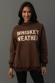 A smiling woman with long dark hair models the Panache Apparel Collection’s Brown Whiskey Weather & Bourbon Weather Reversible Sweatshirt, featuring bold white “WHISKEY WEATHER” lettering, against a plain gray background.