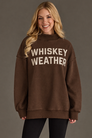 A smiling woman with long blonde hair wears the Panache Apparel Collection’s Brown Whiskey Weather & Bourbon Weather Reversible Sweatshirt, featuring “WHISKEY WEATHER” in white letters, paired with black pants and standing against a plain gray background.