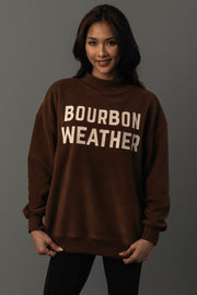A woman is wearing the Panache Apparel Collection’s Brown Whiskey Weather & Bourbon Weather Reversible Sweatshirt, featuring “BOURBON WEATHER” printed in large white letters, standing against a plain gray background and smiling slightly.