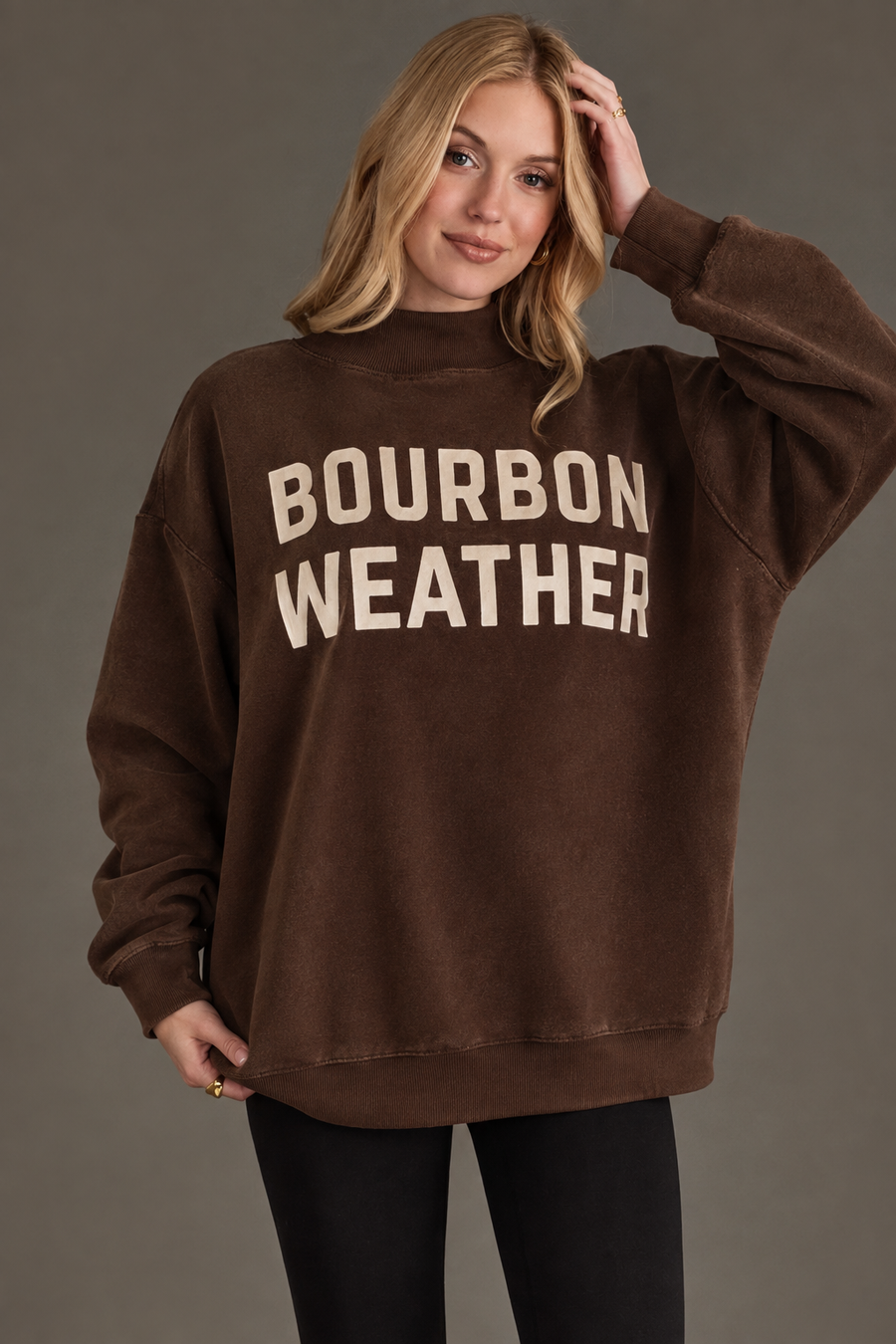 A woman wearing the Panache Apparel Collection’s brown Whiskey Weather & Bourbon Weather reversible sweatshirt stands against a gray background, smiling and touching her hair.