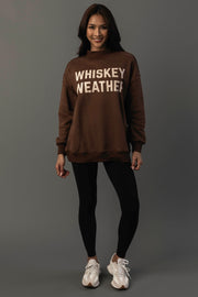 A person stands smiling against a plain gray background, wearing the Panache Apparel Collection’s Brown Whiskey Weather & Bourbon Weather Reversible Sweatshirt, paired with black leggings and white sneakers.