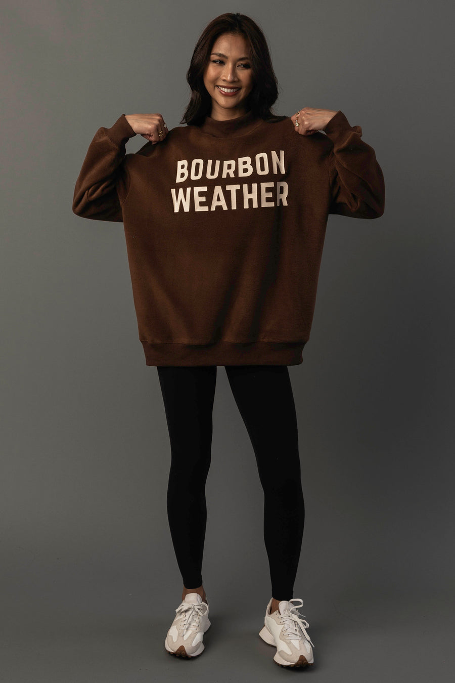 A smiling woman stands against a gray background, holding the sides of her Panache Apparel Collection Brown Whiskey Weather & Bourbon Weather Reversible Sweatshirt, which features “BOURBON WEATHER” printed in bold white letters; she pairs it with black leggings and white sneakers.