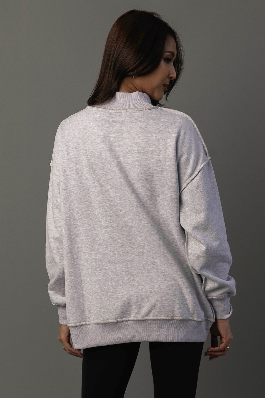 A person with long brown hair, wearing the Light Gray Whiskey Weather & Bourbon Weather Reversible Sweatshirt from Panache Apparel Collection and black pants, stands facing away from the camera against a plain gray background.