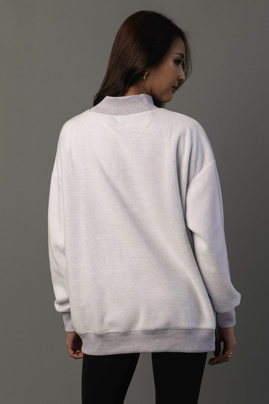 A woman with long brown hair stands facing away, wearing the Panache Apparel Collection’s Light Gray Whiskey Weather & Bourbon Weather Reversible Sweatshirt with black pants against a plain gray background.