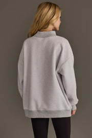 A woman with long blonde hair, seen from behind, wears the Panache Apparel Collection’s Light Gray Whiskey Weather & Bourbon Weather Reversible Sweatshirt paired with black leggings, standing against a plain, neutral background.
