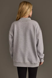 A person with long blonde hair, seen from behind, is wearing the Light Gray Whiskey Weather & Bourbon Weather Reversible Sweatshirt by Panache Apparel Collection and black pants, standing against a plain brown background.