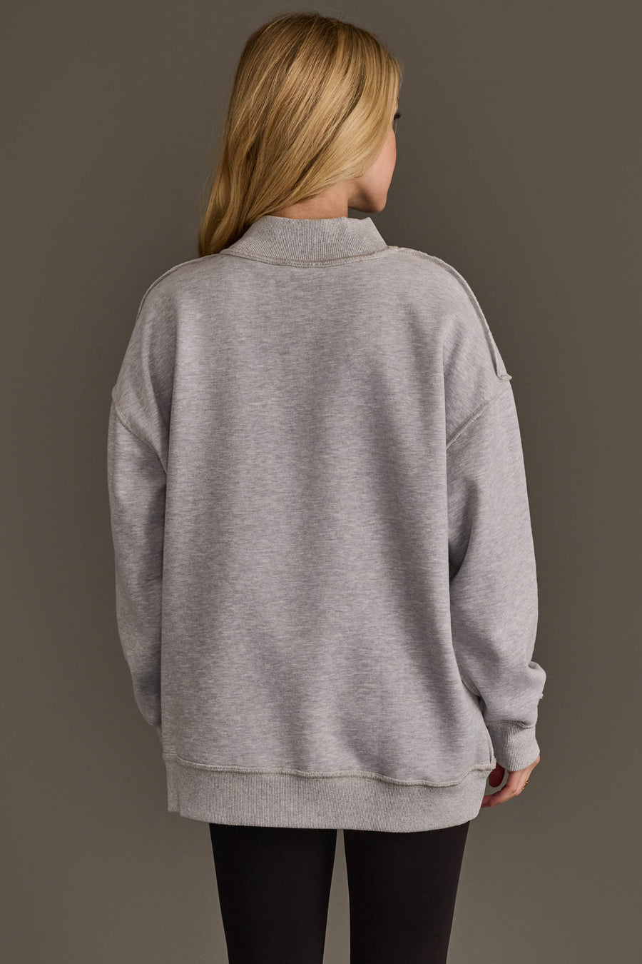 A person with long blonde hair, seen from behind, is wearing the Light Gray Whiskey Weather & Bourbon Weather Reversible Sweatshirt by Panache Apparel Collection and black pants, standing against a plain brown background.
