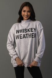 A woman with long dark hair is smiling at the camera while wearing the Panache Apparel Collection’s Light Gray Whiskey Weather & Bourbon Weather Reversible Sweatshirt, featuring bold black lettering, and standing against a plain gray background.