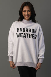 A woman smiles, standing against a gray background, wearing the Panache Apparel Collection’s Light Gray Whiskey Weather & Bourbon Weather Reversible Sweatshirt with ‘BOURBON WEATHER’ in bold black letters across the front.