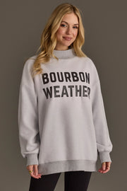 A woman with long blonde hair wears the Panache Apparel Collection Light Gray Whiskey Weather & Bourbon Weather Reversible Sweatshirt, featuring “BOURBON WEATHER” in bold black letters, as she stands against a plain background.