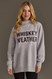 A woman with long blonde hair wears the Panache Apparel Collection Light Gray Whiskey Weather & Bourbon Weather Reversible Sweatshirt, featuring bold black WHISKEY WEATHER lettering, while standing against a plain background.