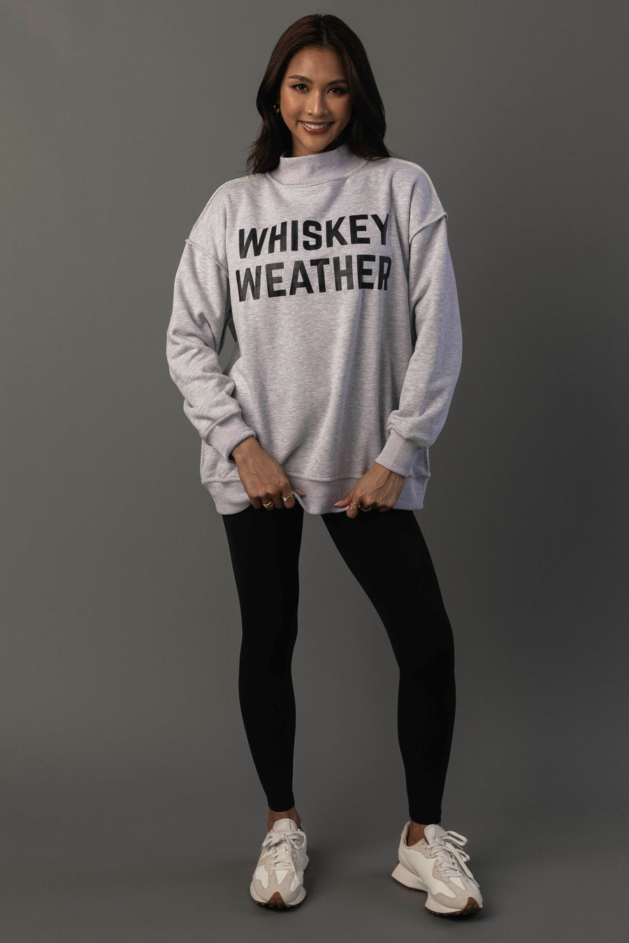 A woman stands against a plain background, smiling, wearing the Panache Apparel Collection Light Gray Whiskey Weather & Bourbon Weather Reversible Sweatshirt, paired with black leggings and white sneakers.