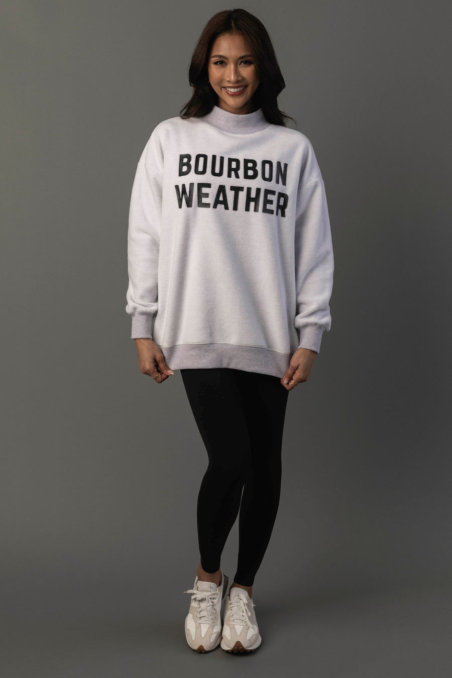 A smiling person wears the Panache Apparel Collection Light Gray Whiskey Weather & Bourbon Weather Reversible Sweatshirt, paired with black leggings and white sneakers, standing against a plain gray background.