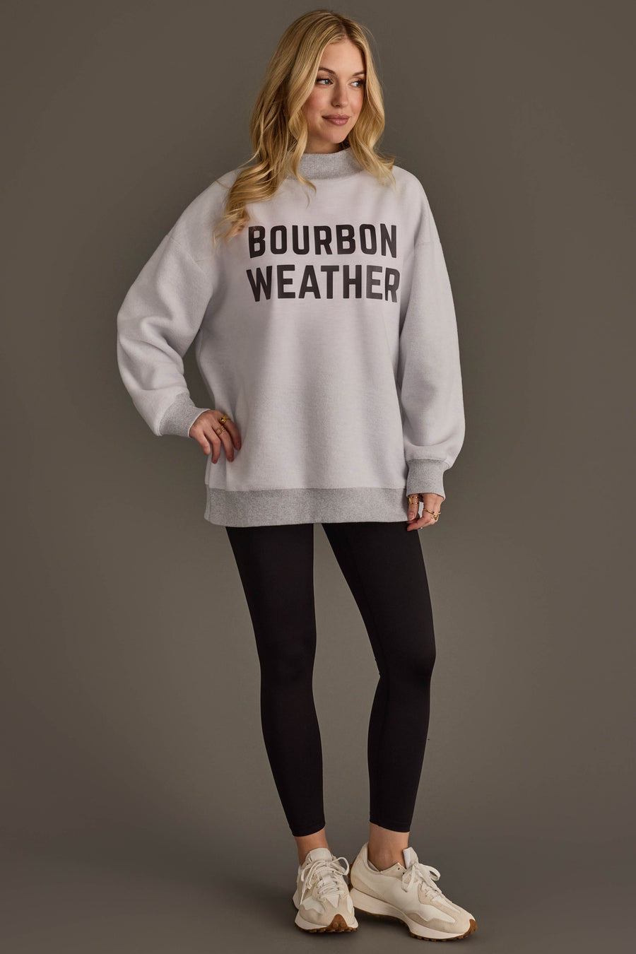A woman is wearing a Light Gray Whiskey Weather & Bourbon Weather Reversible Sweatshirt from Panache Apparel Collection, paired with black leggings and white sneakers, standing against a plain background and smiling.