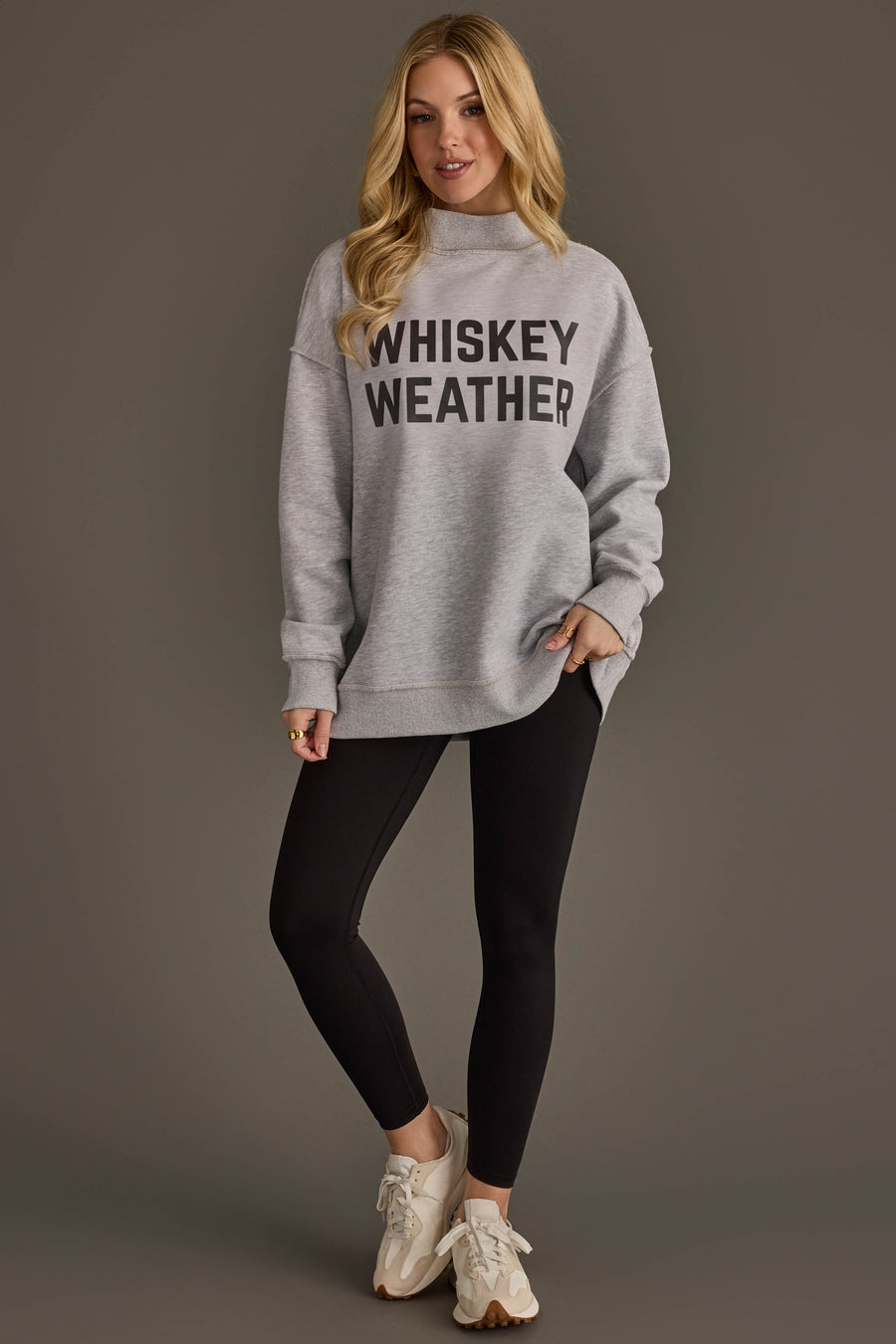 A woman with long blonde hair wears the Panache Apparel Collection Light Gray Whiskey Weather & Bourbon Weather Reversible Sweatshirt, paired with black leggings and white sneakers, standing against a plain brown background.