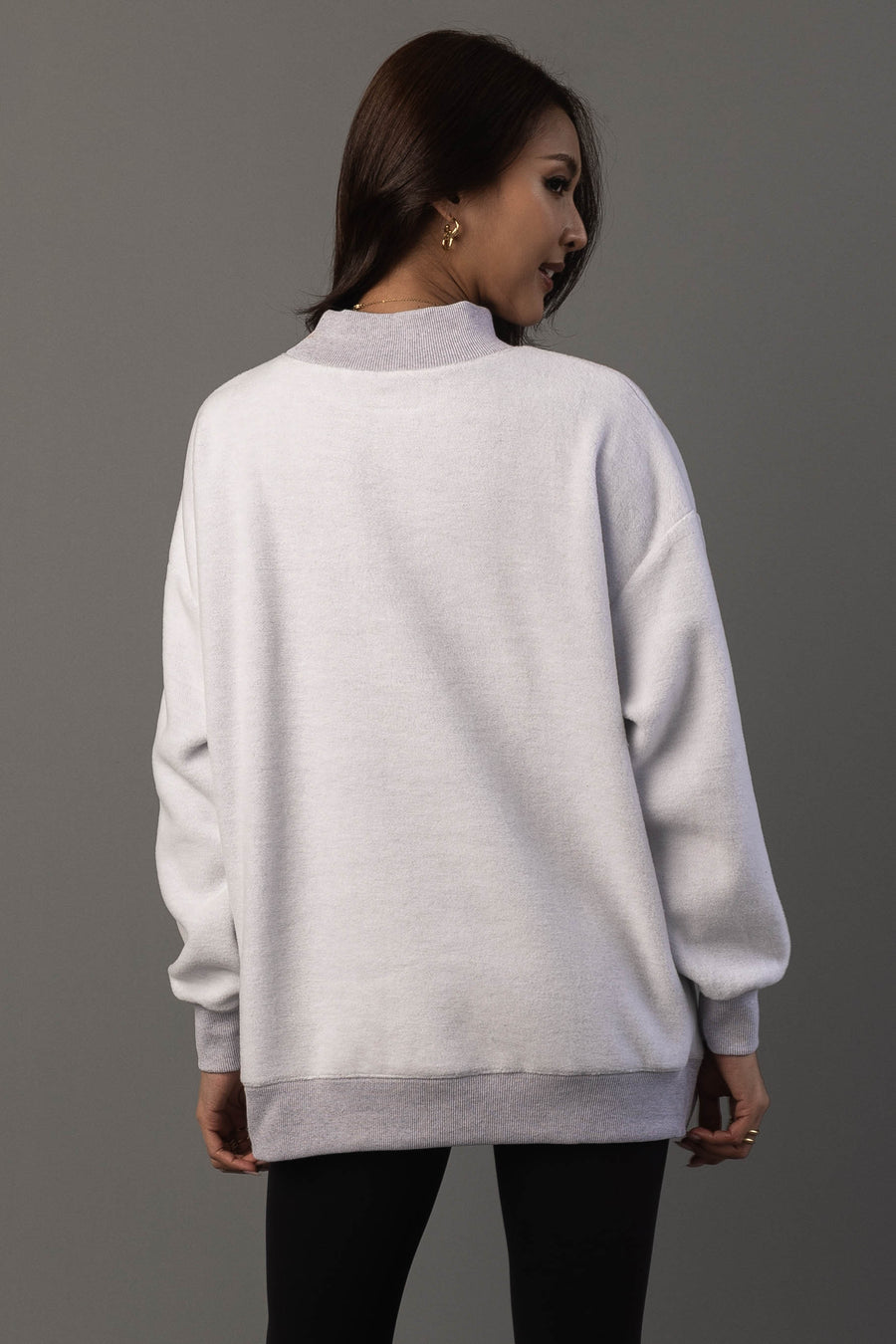 A woman with long brown hair, wearing gold hoop earrings and black leggings, stands with her back to the camera against a gray background, modeling the Panache Apparel Collection’s Light Gray Day Drinking & Hungover Reversible Sweatshirt.