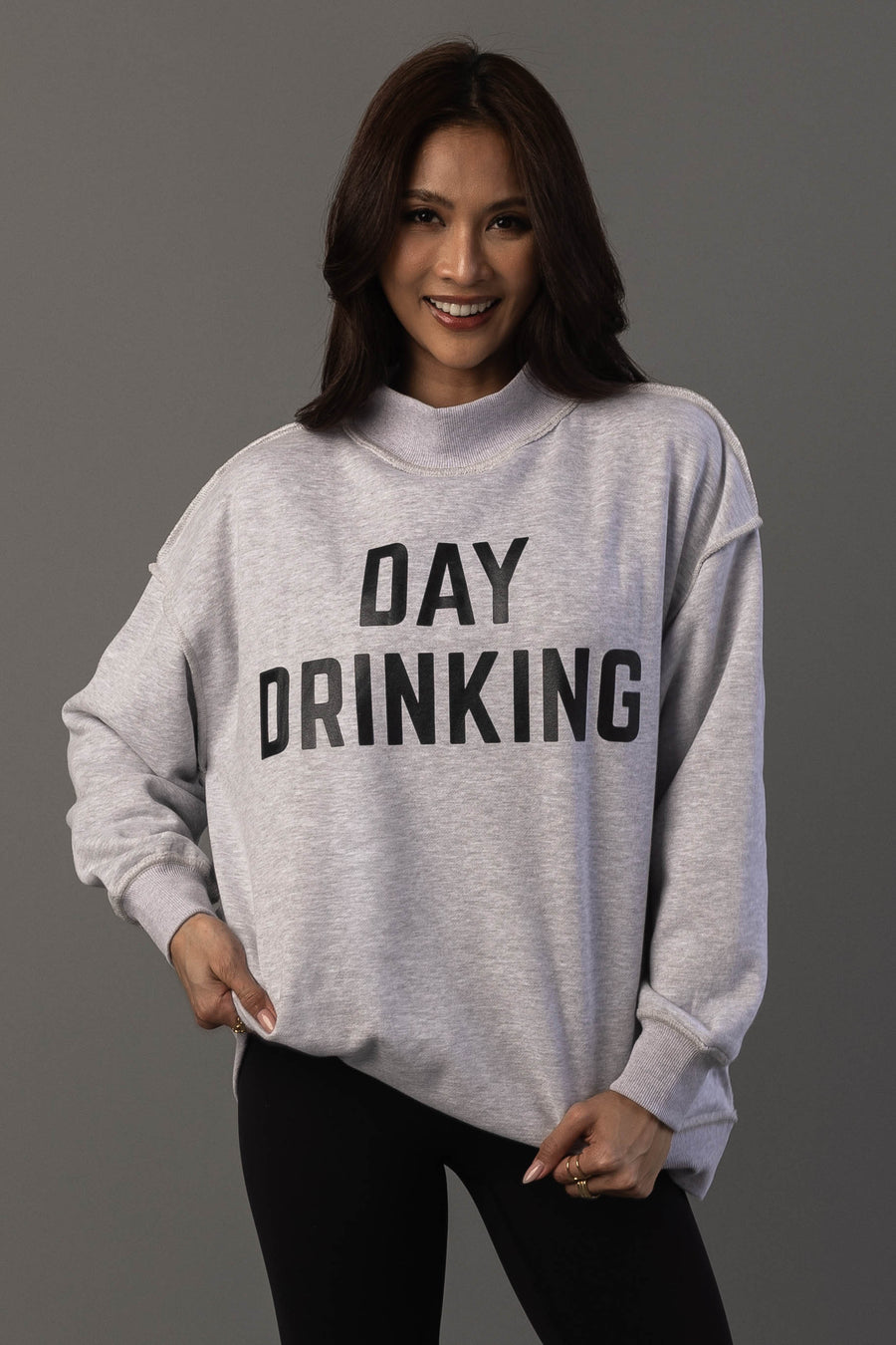A woman smiles while wearing the Panache Apparel Collection’s Light Gray Day Drinking & Hungover Reversible Sweatshirt, featuring bold black DAY DRINKING lettering, and stands against a plain gray background.