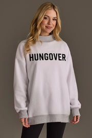 A woman with long blonde hair wears the Panache Apparel Collection Light Gray Day Drinking & Hungover Reversible Sweatshirt, styled in a loose, oversized fit with bold black lettering, paired with black leggings and multiple rings, standing against a plain brown background.