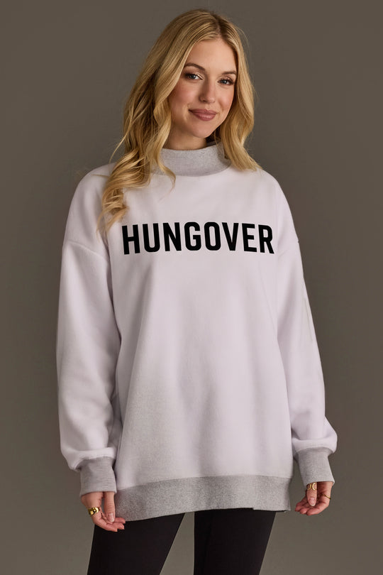 A woman with long blonde hair wears the Panache Apparel Collection Light Gray Day Drinking & Hungover Reversible Sweatshirt, styled in a loose, oversized fit with bold black lettering, paired with black leggings and multiple rings, standing against a plain brown background.