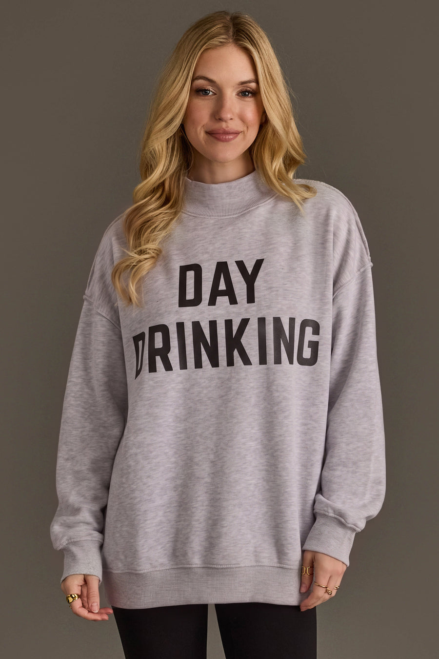 A smiling woman with long blonde hair wears the Panache Apparel Collection Light Gray Day Drinking & Hungover Reversible Sweatshirt, featuring an oversized fit and bold black “DAY DRINKING” lettering. She stands against a plain, dark background.
