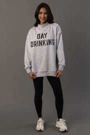 A woman stands against a gray background, smiling with her hands by her sides, wearing the Panache Apparel Collection’s Light Gray Day Drinking & Hungover Reversible Sweatshirt, paired with black leggings and white sneakers.