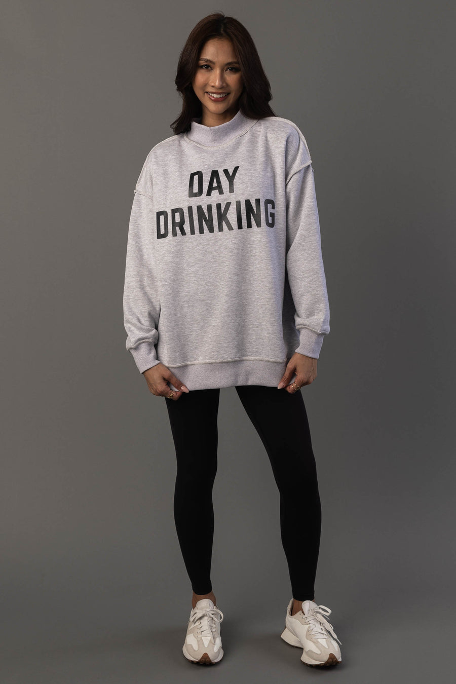 A woman stands against a gray background, smiling with her hands by her sides, wearing the Panache Apparel Collection’s Light Gray Day Drinking & Hungover Reversible Sweatshirt, paired with black leggings and white sneakers.