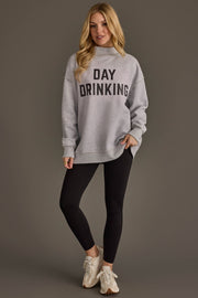 A woman with long blonde hair wears the Panache Apparel Collection Light Gray Day Drinking & Hungover Reversible Sweatshirt, styled in an oversized fit, paired with black leggings and white sneakers, standing against a plain gray background.