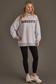 A woman stands against a plain background, wearing the Panache Apparel Collection’s Light Gray Day Drinking & Hungover Reversible Sweatshirt in an oversized fit, paired with black leggings and white sneakers. Her blonde hair frames her face as she smiles slightly, and the cozy sweatshirt perfectly highlights her laid-back vibe.