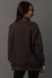A woman with long dark hair, dressed in the Panache Apparel Collection’s Dark Gray Whiskey Weather & Bourbon Weather Reversible Sweatshirt and black pants, stands turned slightly away from the camera against a plain gray background.