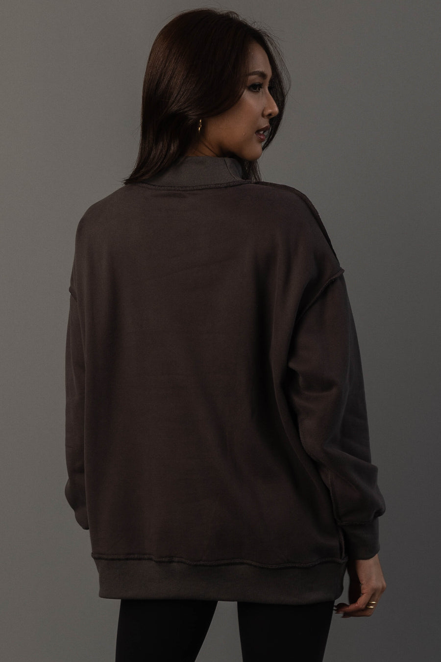A woman with long dark hair, dressed in the Panache Apparel Collection’s Dark Gray Whiskey Weather & Bourbon Weather Reversible Sweatshirt and black pants, stands turned slightly away from the camera against a plain gray background.