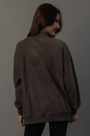 A woman with long brown hair is wearing the Panache Apparel Collection’s Dark Gray Whiskey Weather & Bourbon Weather Reversible Sweatshirt and black pants, standing against a gray background, facing away from the camera and looking over her shoulder with a slight smile.