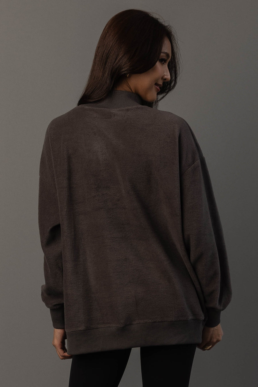 A woman with long brown hair is wearing the Panache Apparel Collection’s Dark Gray Whiskey Weather & Bourbon Weather Reversible Sweatshirt and black pants, standing against a gray background, facing away from the camera and looking over her shoulder with a slight smile.
