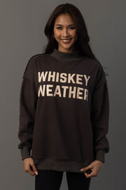 A person with long dark hair smiles while wearing the Panache Apparel Collection’s Dark Gray Whiskey Weather & Bourbon Weather Reversible Sweatshirt, featuring “WHISKEY WEATHER” in large white letters. The background is plain gray.