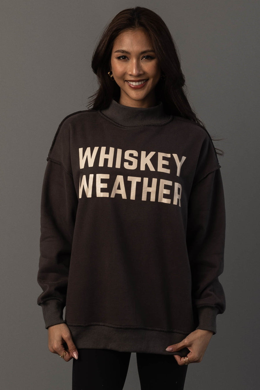 A person with long dark hair smiles while wearing the Panache Apparel Collection’s Dark Gray Whiskey Weather & Bourbon Weather Reversible Sweatshirt, featuring “WHISKEY WEATHER” in large white letters. The background is plain gray.