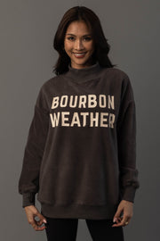 A woman with long dark hair is wearing the Panache Apparel Collection’s Dark Gray Whiskey Weather & Bourbon Weather Reversible Sweatshirt, featuring “BOURBON WEATHER” in large white letters. She stands against a plain gray background, smiling at the camera.