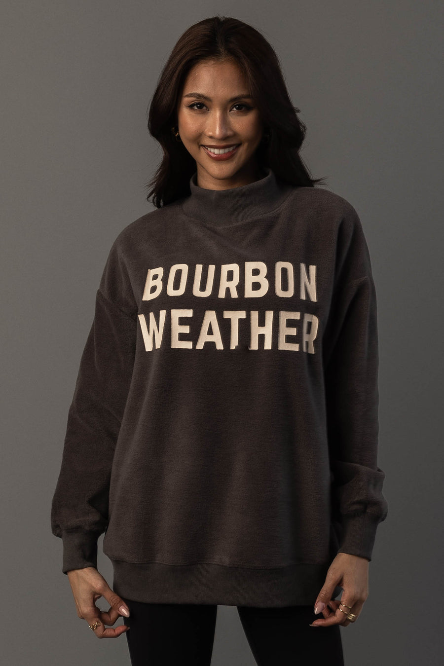 A woman with long dark hair is wearing the Panache Apparel Collection’s Dark Gray Whiskey Weather & Bourbon Weather Reversible Sweatshirt, featuring “BOURBON WEATHER” in large white letters. She stands against a plain gray background, smiling at the camera.
